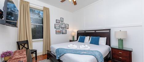 A hotel room with a bed, a TV, a ceiling fan, a window with curtains, a bedside table with a lamp, and a small table with a remote control.