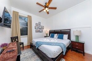 A hotel room with a bed, a TV, a ceiling fan, a window with curtains, a bedside table with a lamp, and a small table with a remote control. - 30A Inn & Suites (Santa Rosa Beach)