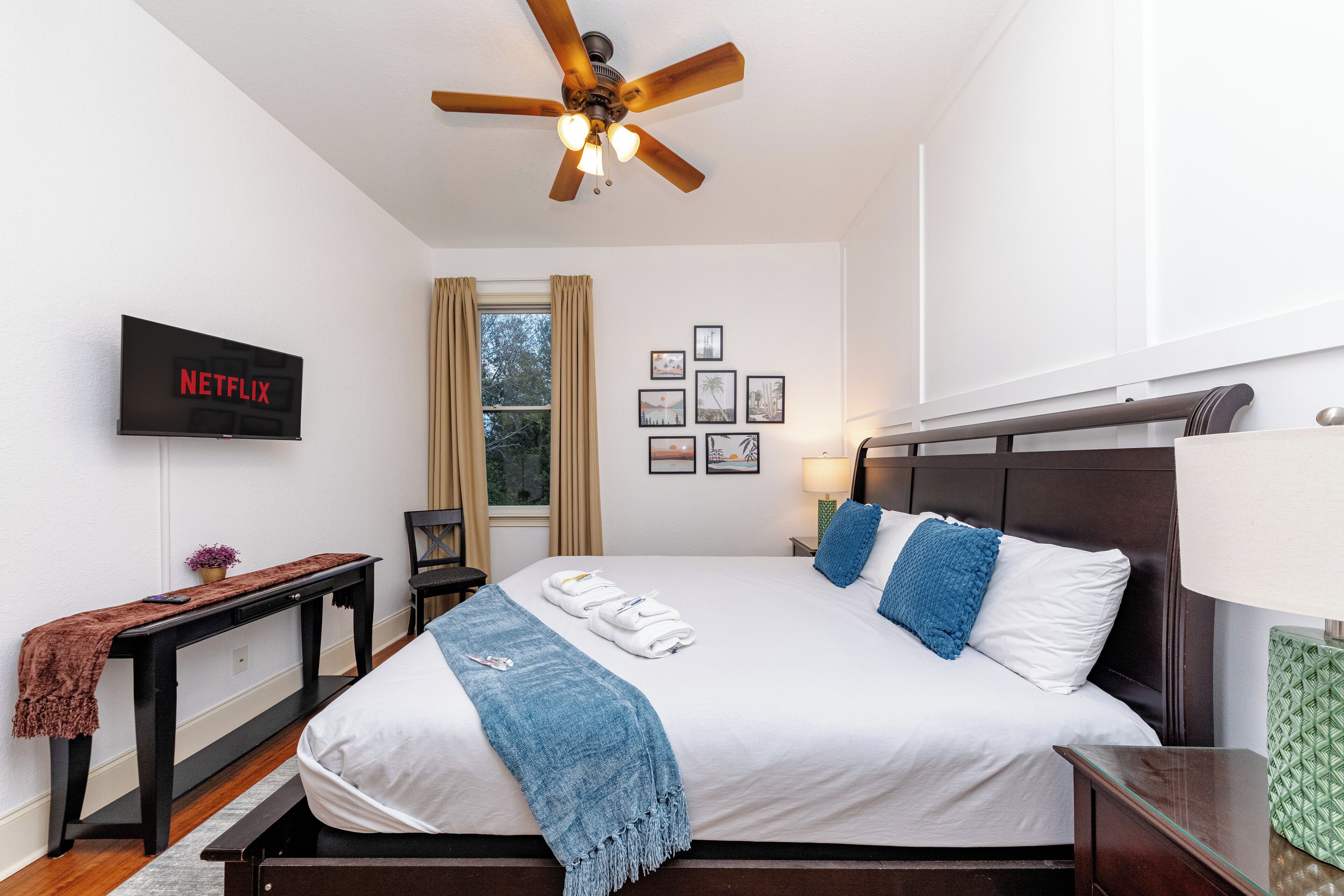 A hotel room with a bed, a ceiling fan, a TV displaying 'NETFLIX', a window with curtains, and framed pictures on the wall.