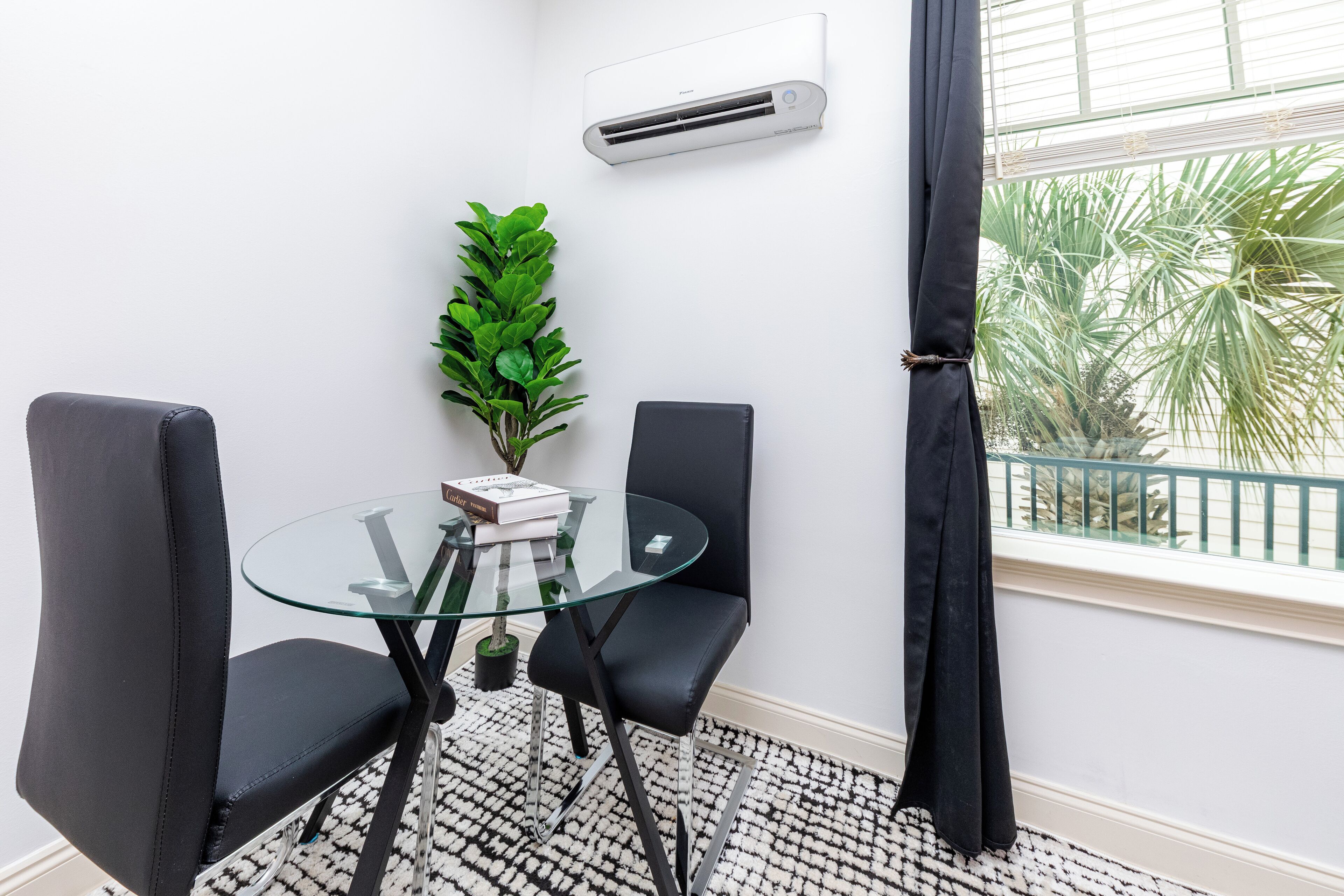 Una habitación con una mesa de vidrio, dos sillas negras, una planta en maceta, un aire acondicionado blanco y una ventana con vista a palmeras.