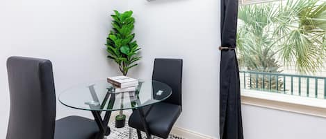A room with a glass table, two black chairs, a potted plant, a white air conditioner, and a window with a view of palm trees.