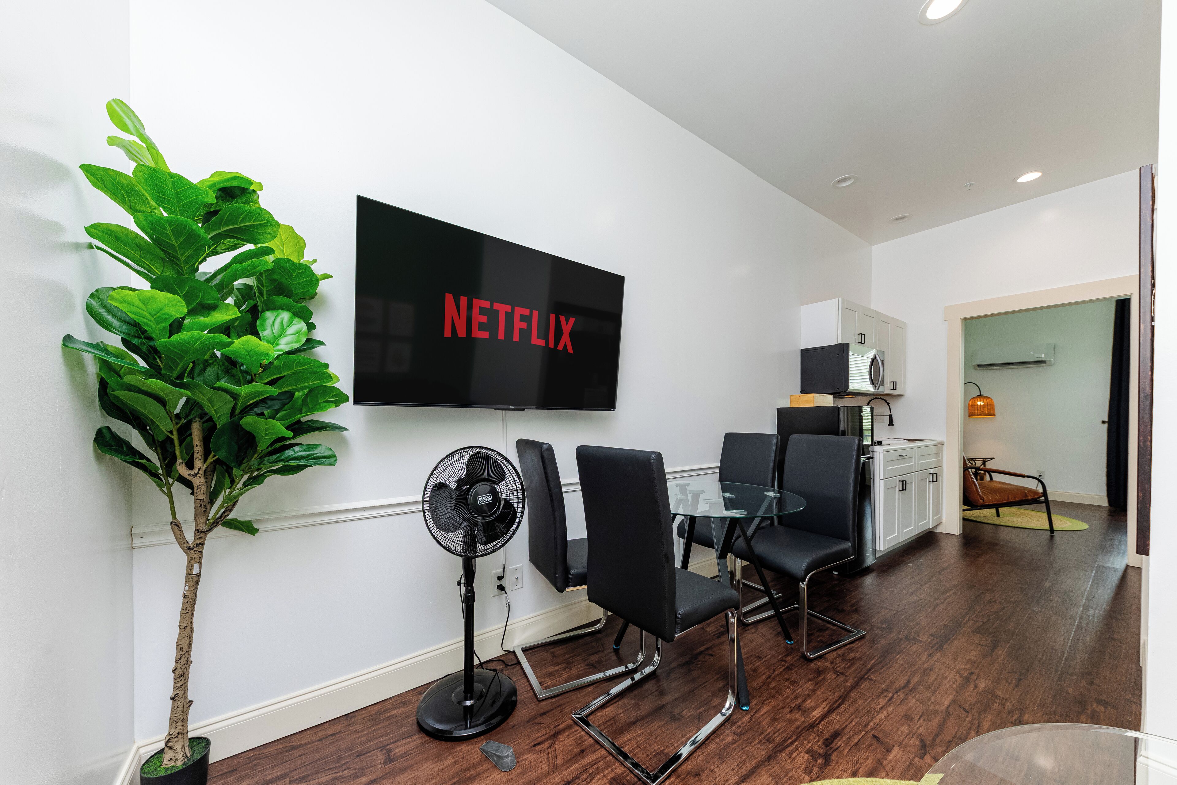 A modern living room with a flat-screen TV, a potted plant, a standing fan, and a dining area with chairs and a table.