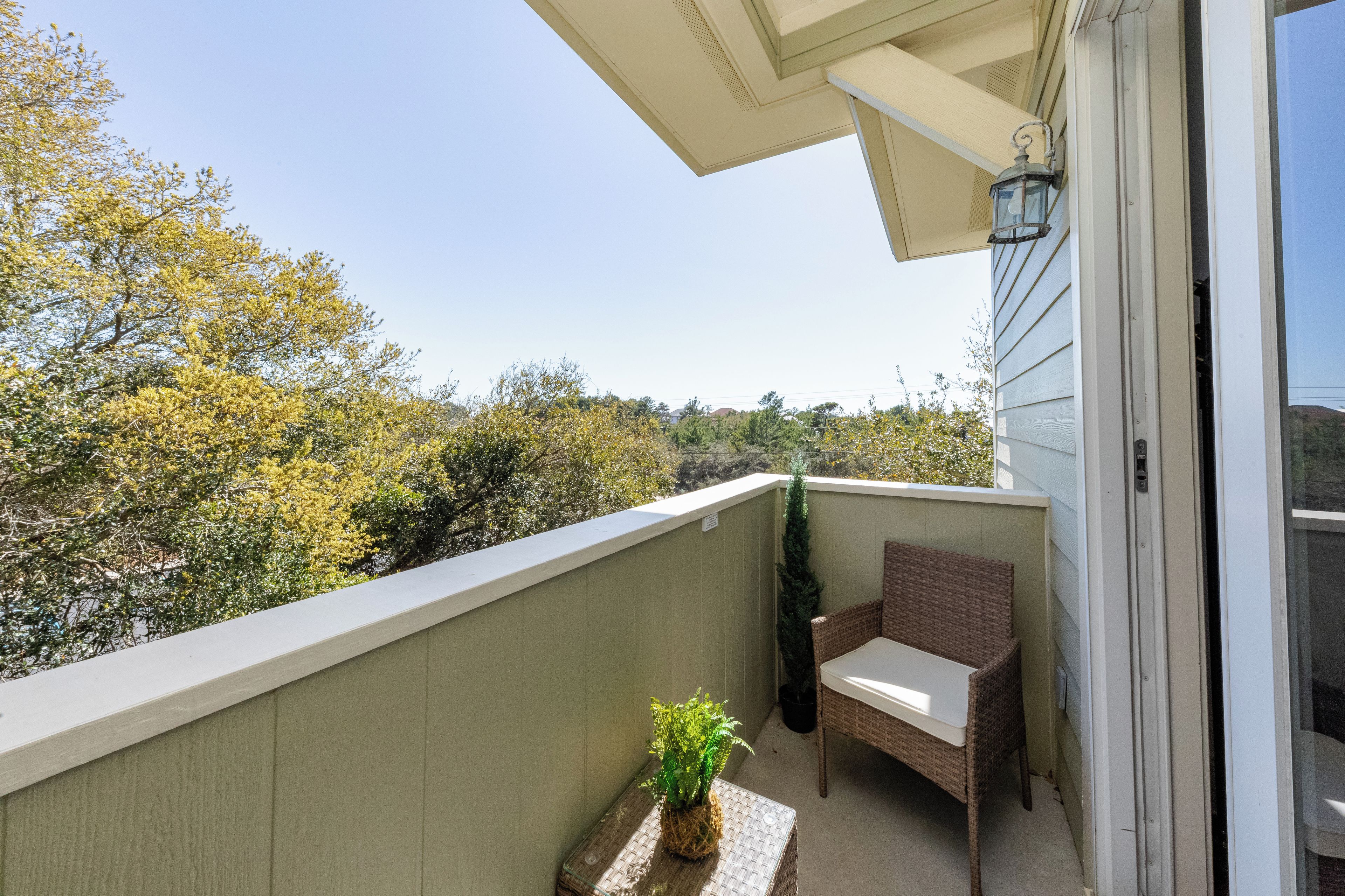 Un balcon avec un fauteuil en osier, une petite table ornée d’une plante, et une vue sur les arbres et les collines.