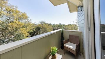 Un balcón con una silla de mimbre, una mesita con una planta y vistas a árboles y colinas.