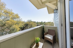 Un balcon avec un fauteuil en osier, une petite table ornée d’une plante, et une vue sur les arbres et les collines.