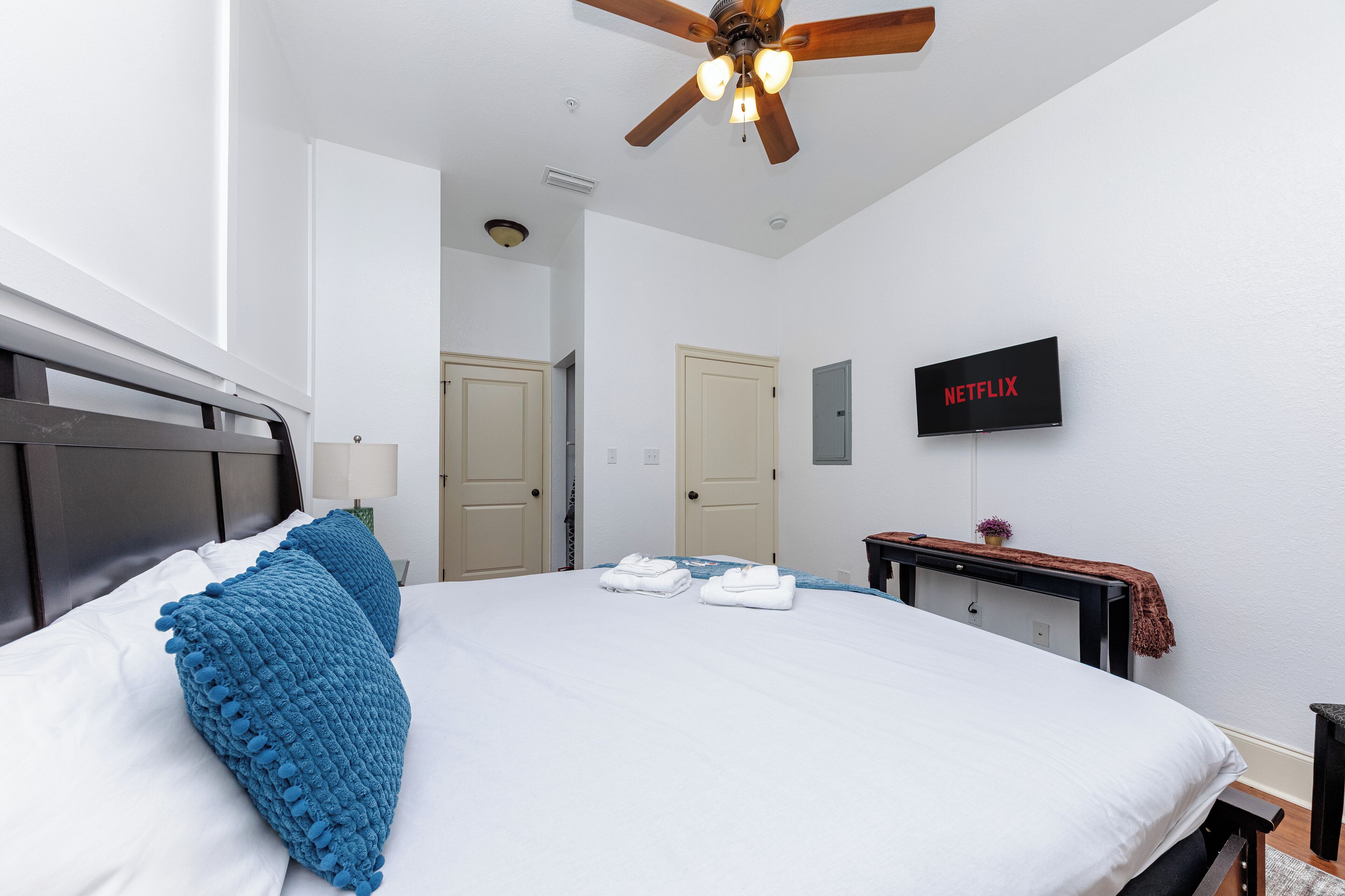 A hotel room with a large bed, a ceiling fan, a TV mounted on the wall, and a desk with a lamp.