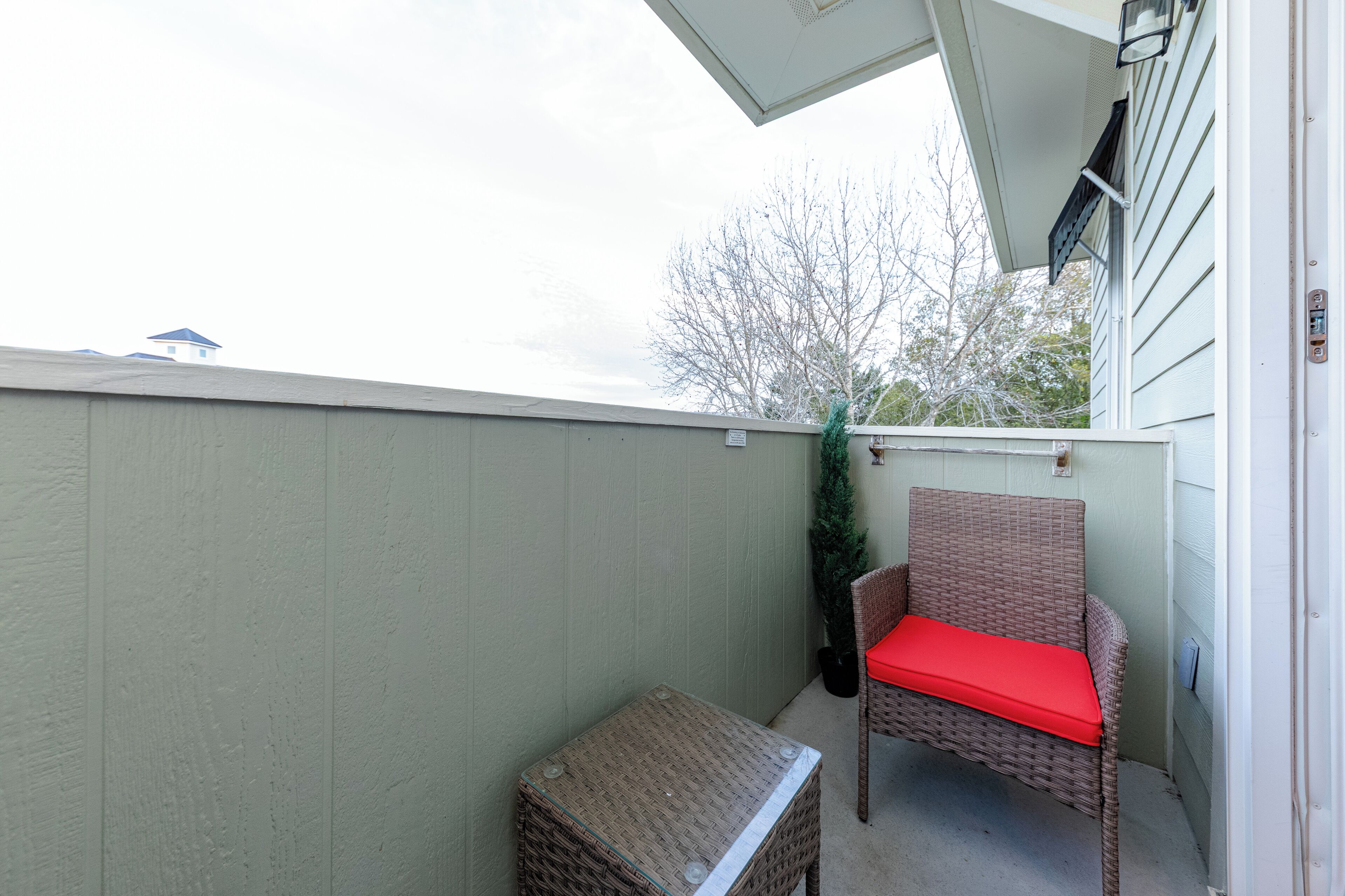 A balcony with a wicker chair and a small table, a green plant, and a view of a building and trees.