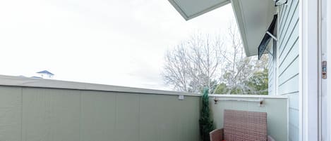 A balcony with a wicker chair and a small table, a green plant, and a view of a building and trees.