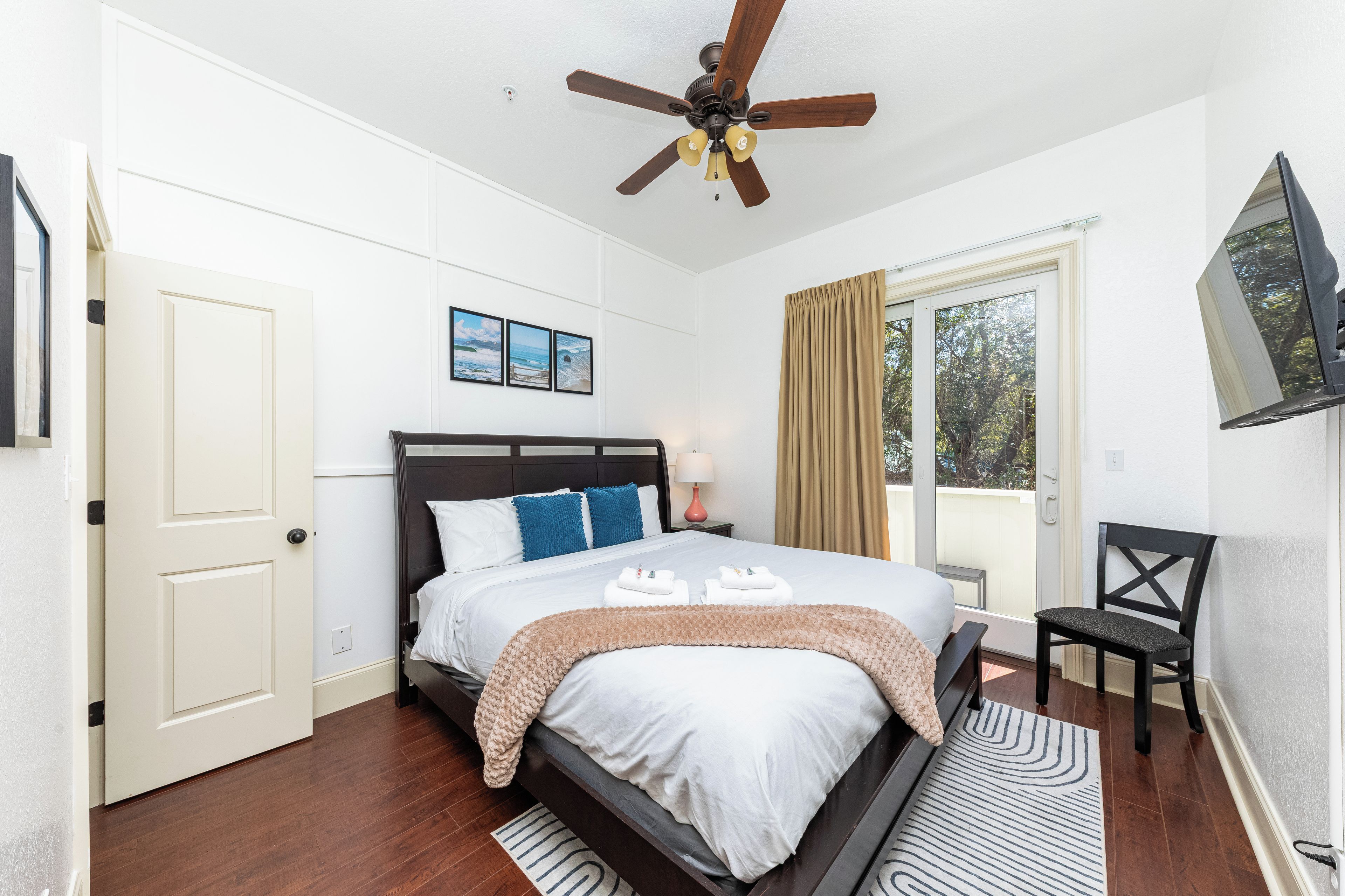 A hotel room with a bed, a ceiling fan, a window with curtains, a TV mounted on the wall, and a door.
