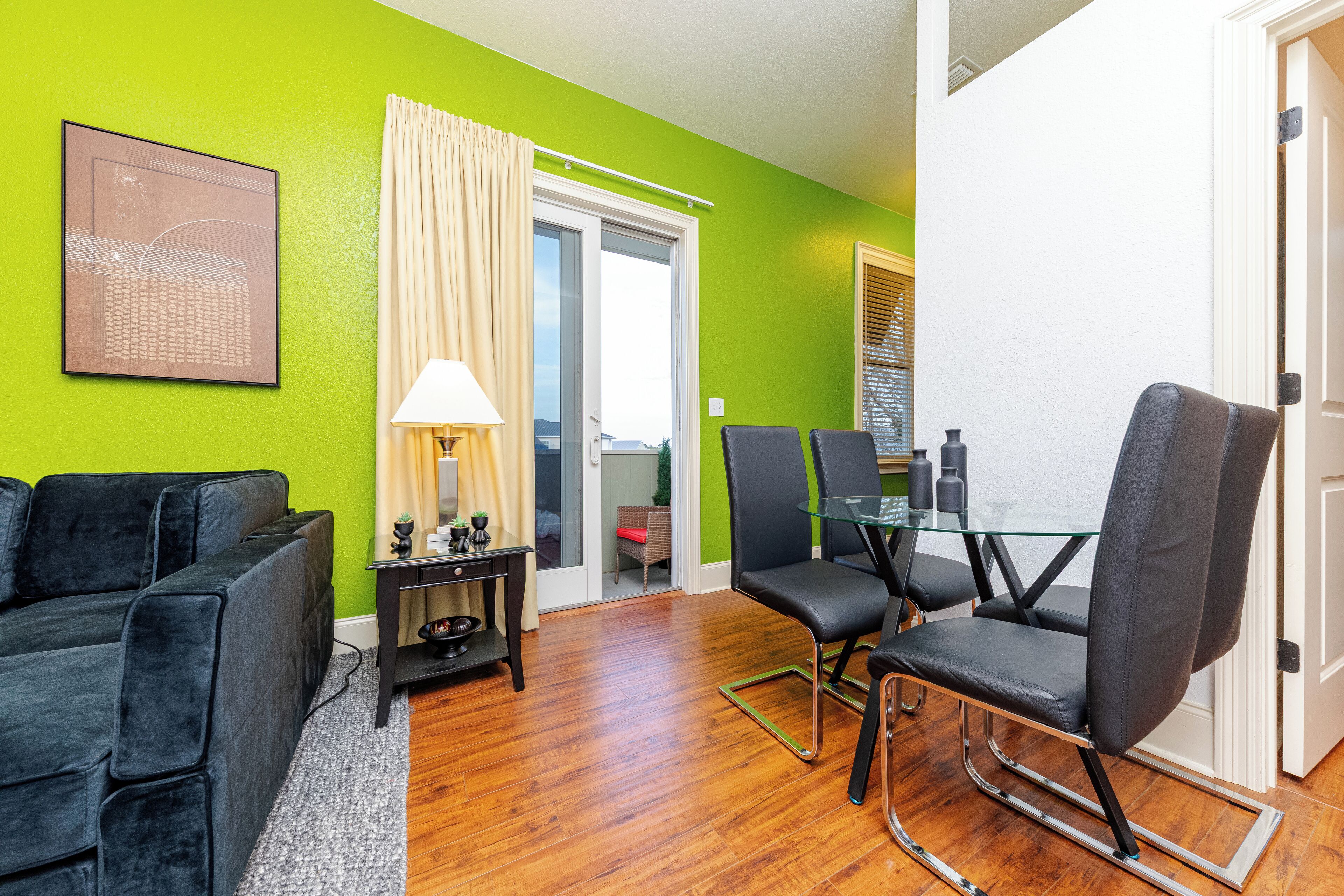A modern living room with a green wall, a black sofa, a glass dining table with chairs, and a sliding glass door leading to a balcony.