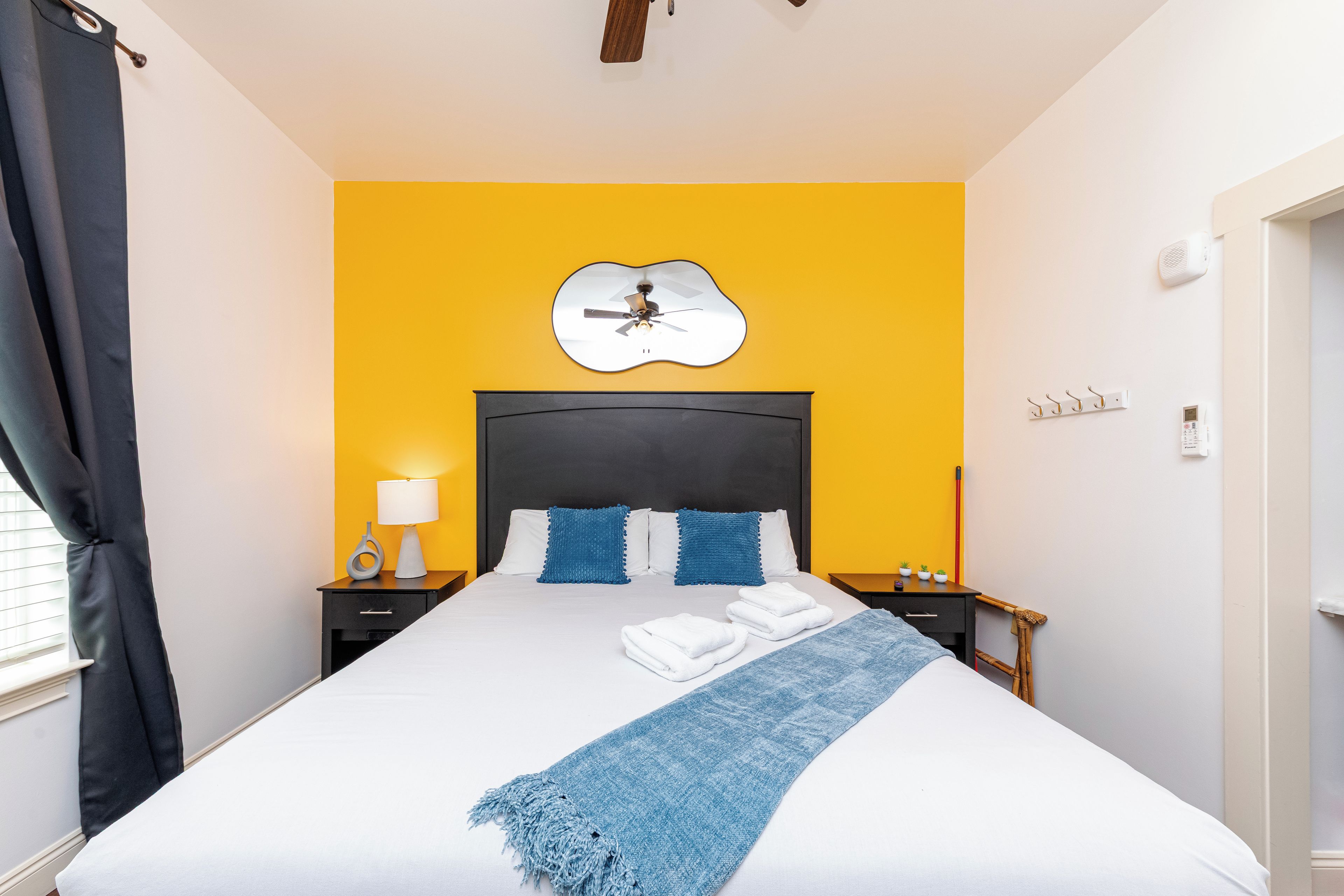 A hotel room with a bed, a nightstand, a lamp, a wall clock, a thermostat, and a window with curtains.