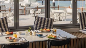 Un comedor con mesas y sillas, vista a barcos y barandilla con vista al agua.