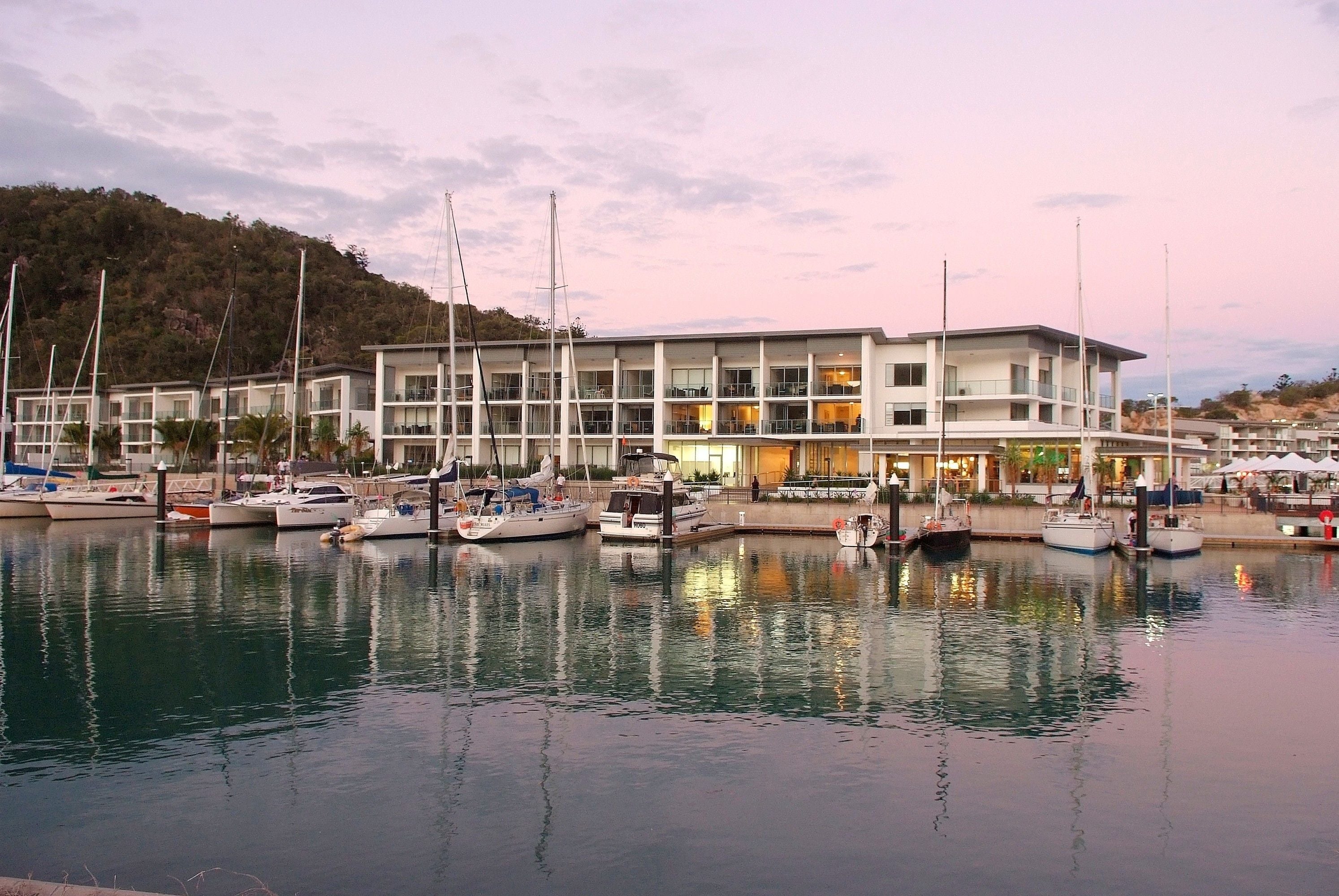 A marina with multiple boats docked, a modern building with balconies, and a hilly landscape in the background.