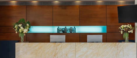 A reception desk with marble top, wooden panel walls, a flat-screen TV, and two flower arrangements.