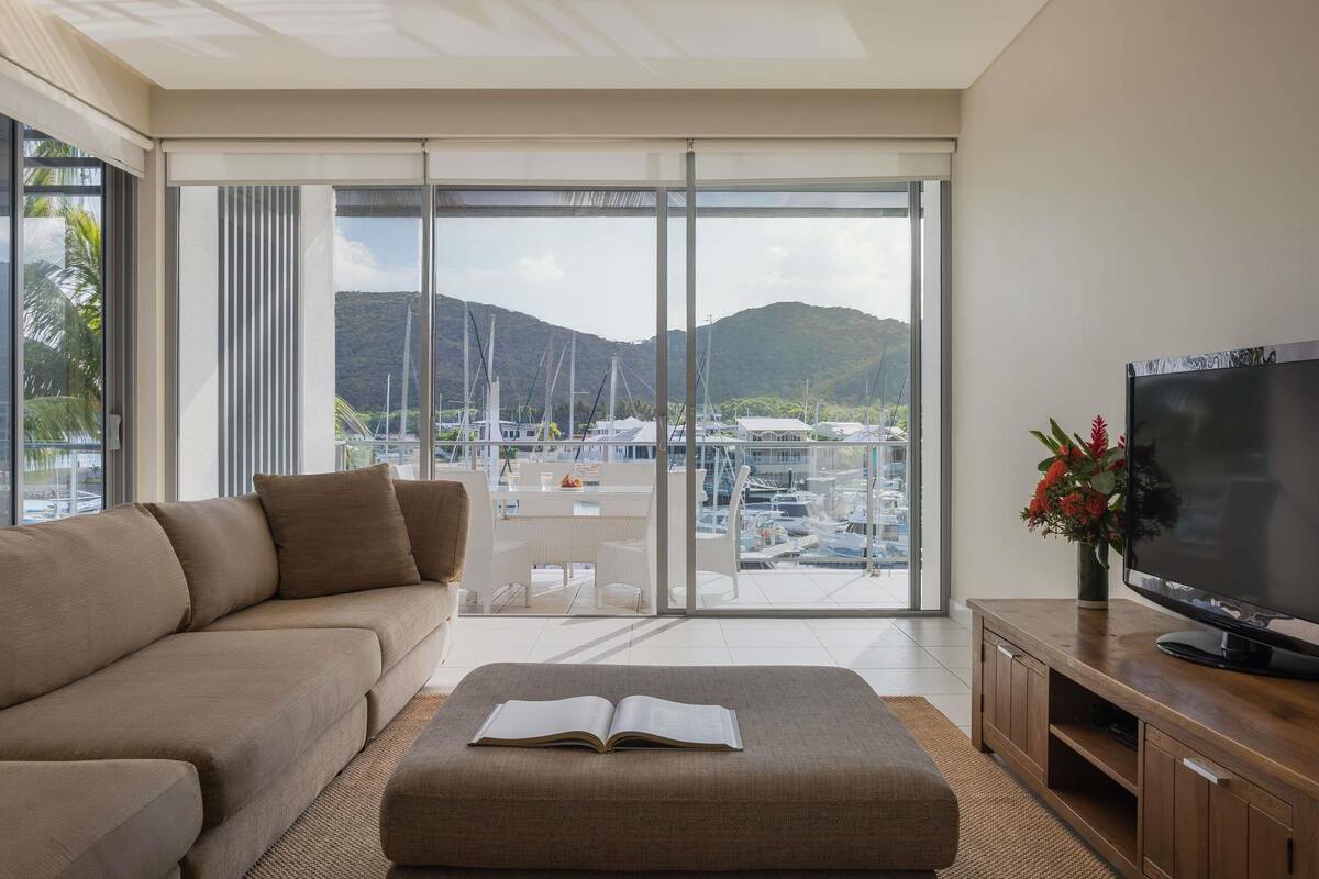Un salon moderne avec un canapé, un pouf, un téléviseur posé sur un meuble en bois, et une vue sur une marina avec des bateaux et des montagnes.