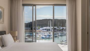 A hotel room with a large bed, a nightstand, a lamp, and a view of a marina with boats and buildings.