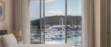 Habitación de hotel con una cama grande, una mesita de noche, una lámpara y vistas a un puerto deportivo con barcos y edificios.