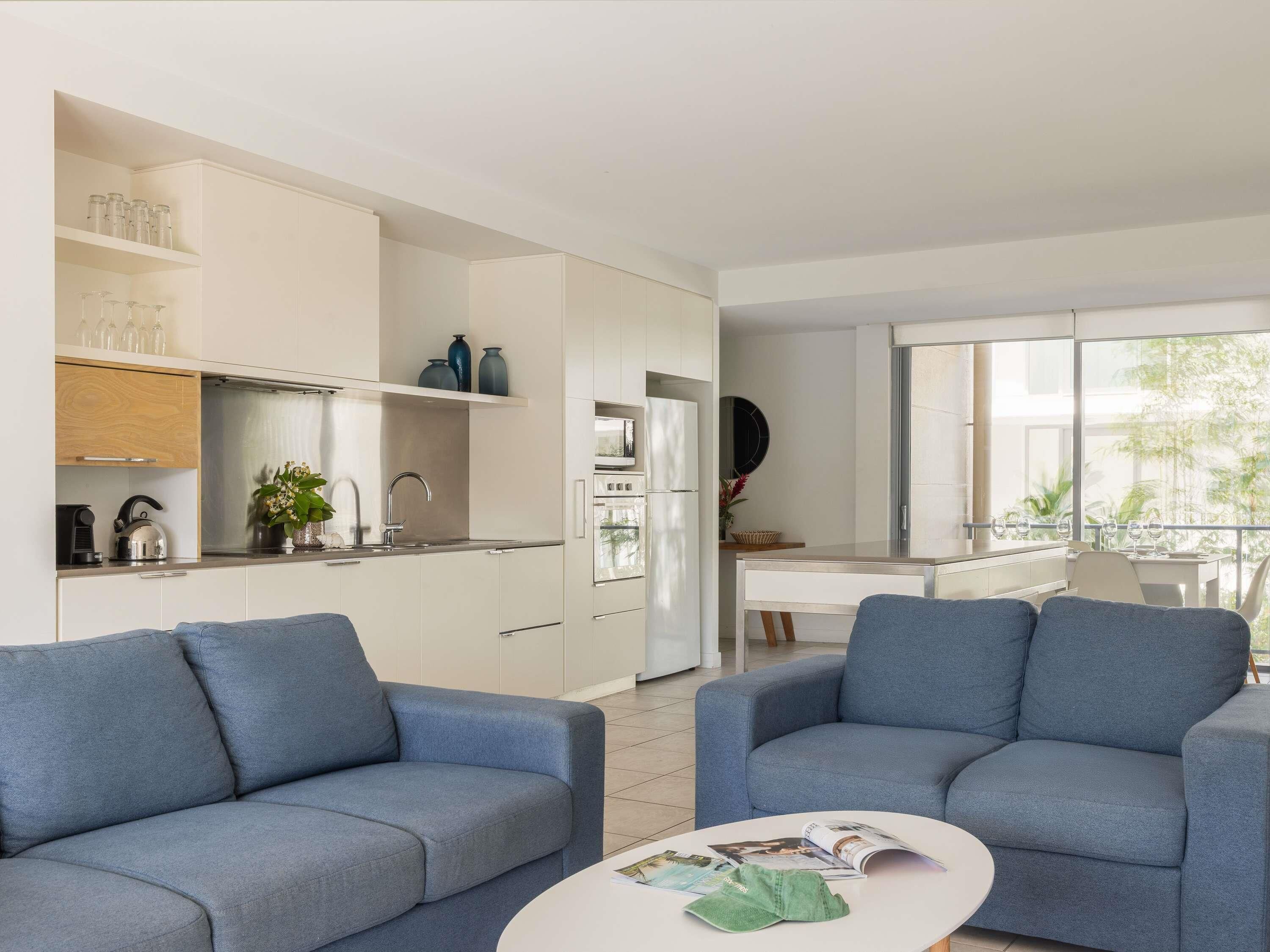 A modern living room with a sofa, a coffee table with magazines, and a kitchen area with cabinets and appliances.