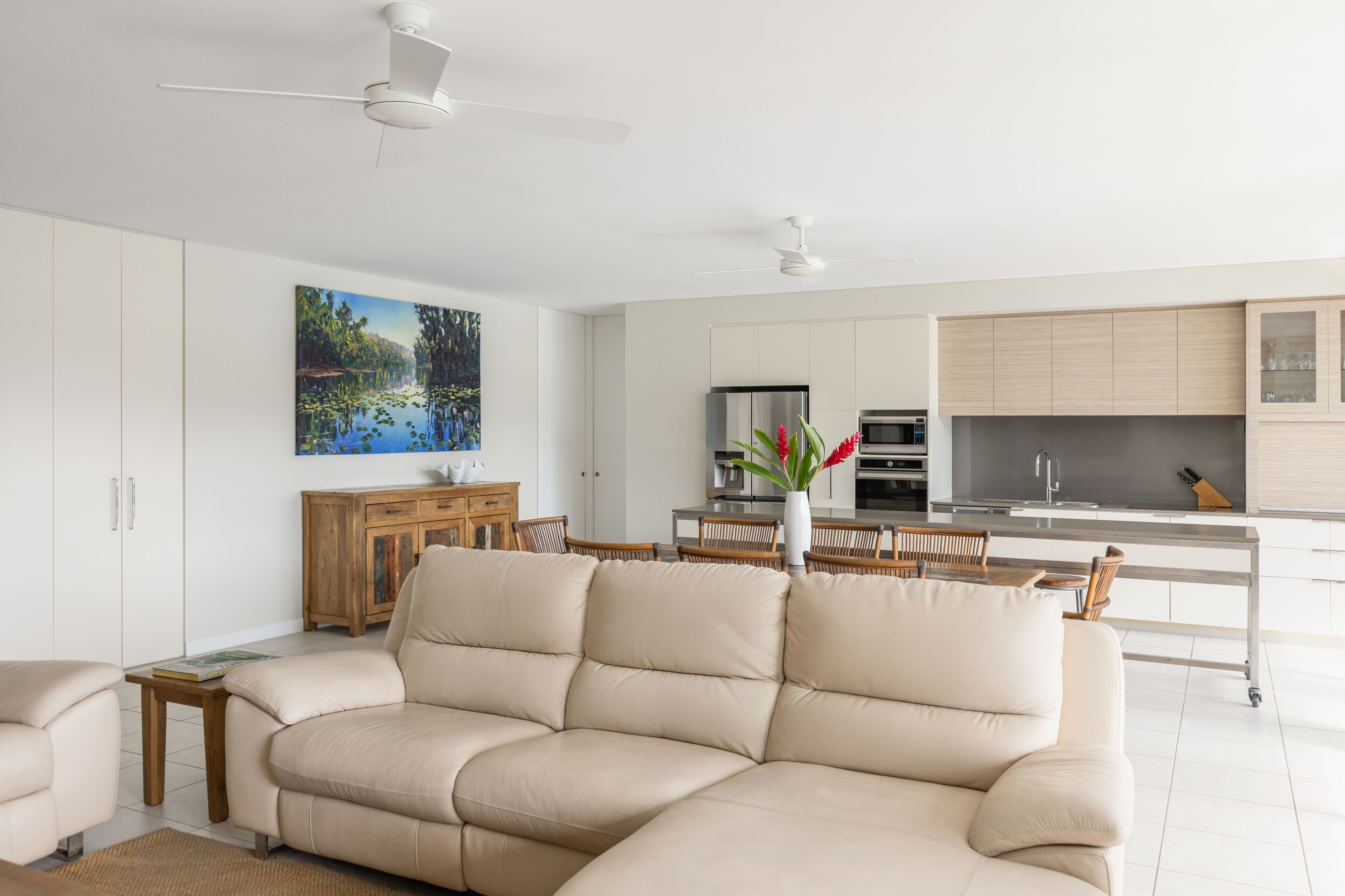 A modern living room with a beige sectional sofa, a wooden cabinet, and a dining area with a table and chairs. There is a kitchen in the background with white cabinets and stainless steel appliances.