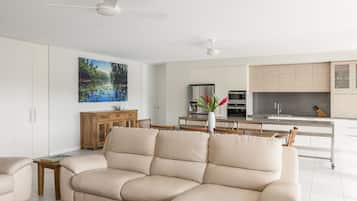 A modern living room with a beige sectional sofa, a wooden cabinet, and a dining area with a table and chairs. There is a kitchen in the background with white cabinets and stainless steel appliances.