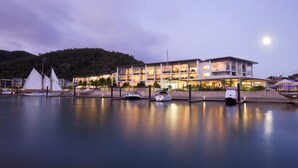 Un hotel frente al agua con varios balcones, un puerto deportivo con barcos y una colina al fondo.