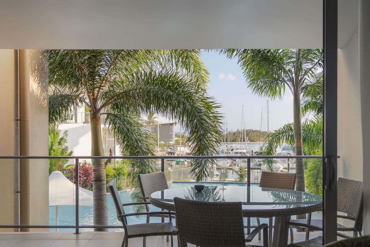 Un balcon doté d’une table et de chaises en verre, offrant une vue sur une marina avec des bateaux et des palmiers.