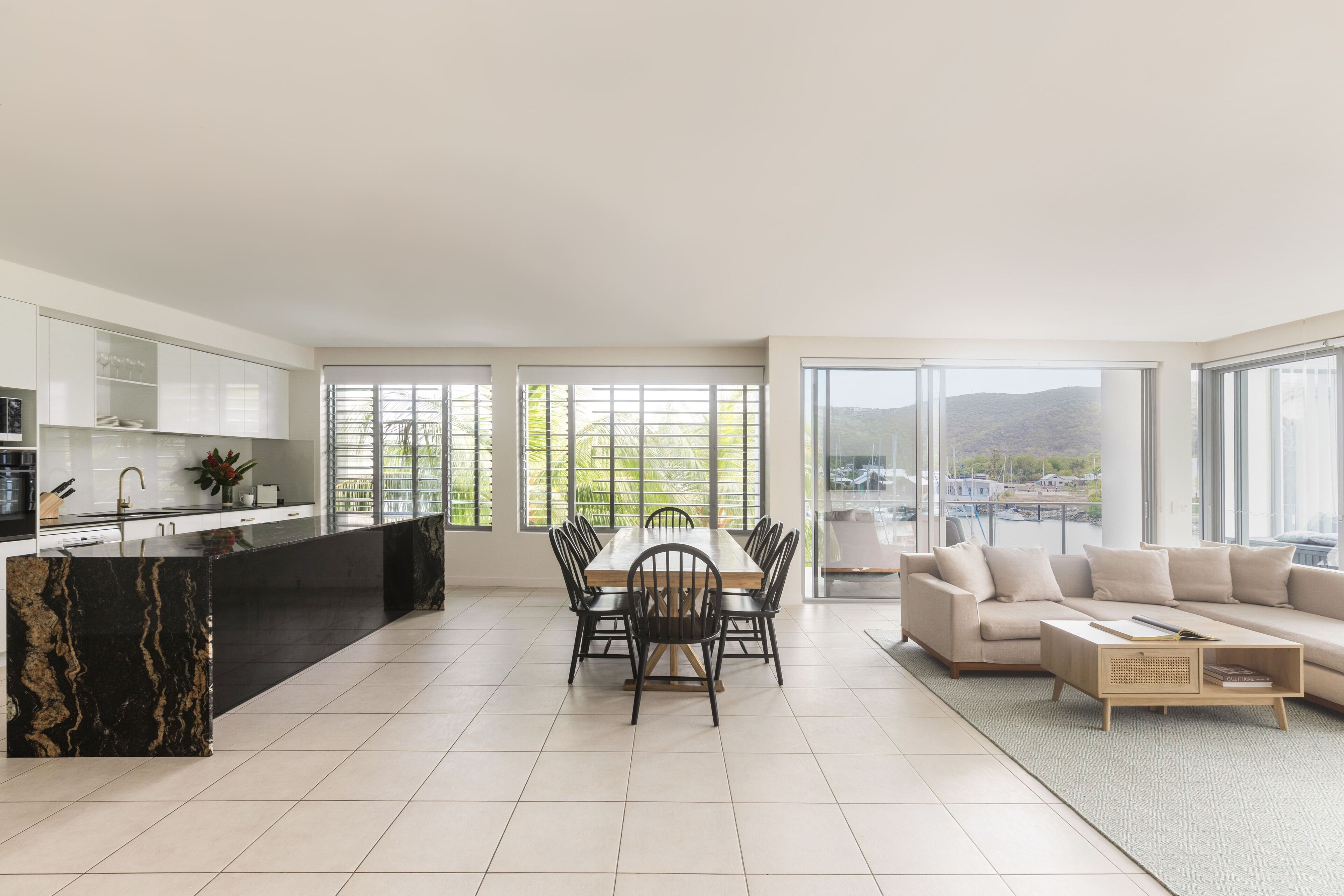 Una cocina moderna con una isla de mármol negro, un comedor con mesa y sillas de madera, un sofá beige y grandes ventanales con vistas a zonas verdes y colinas.