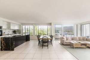 Una cocina moderna con una isla de mármol negro, un comedor con mesa y sillas de madera, un sofá beige y grandes ventanales con vistas a zonas verdes y colinas.