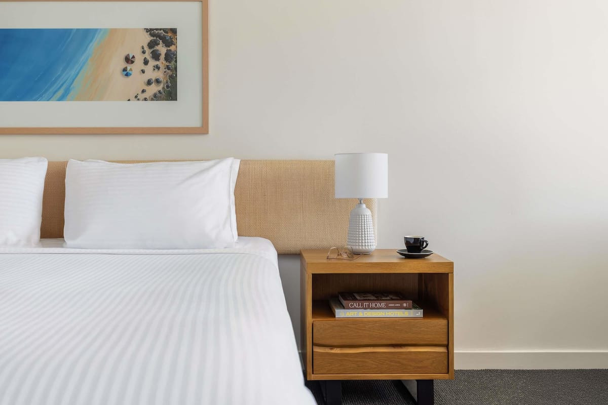 Un lit bien fait, avec du linge de lit blanc, une table de chevet en bois sur laquelle se trouvent une lampe et des livres, et un tableau représentant une scène de plage accroché au mur.