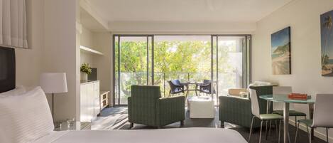 A hotel room with a large bed, a desk with a chair, a small table with chairs, and a balcony with a view of trees and a beach.