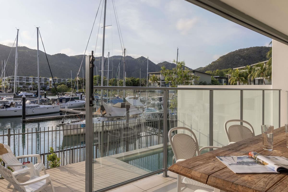 Un balcon donnant sur une marina, une table en bois et une balustrade en verre.