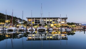 Un puerto deportivo con varias embarcaciones amarradas en aguas tranquilas, un edificio moderno con varios pisos y balcones, y un cielo despejado.