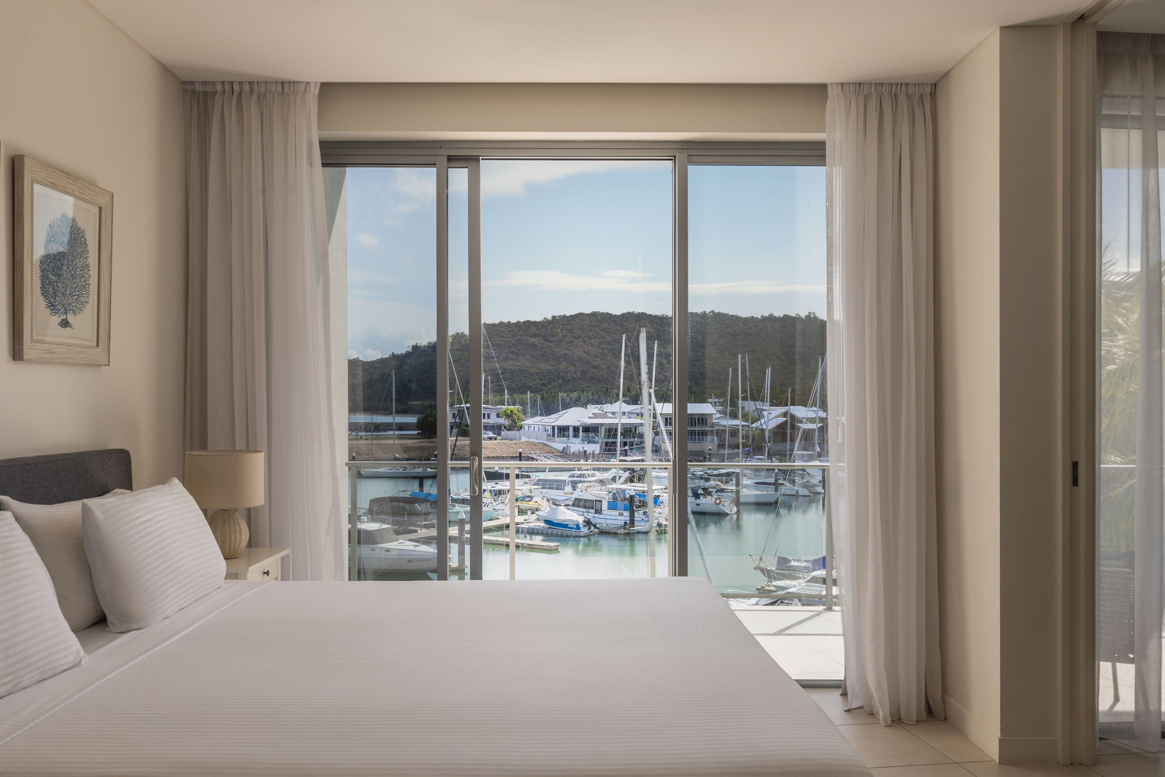 Habitación de hotel con una cama grande, una mesita de noche, una lámpara y vista a un puerto deportivo con barcos.