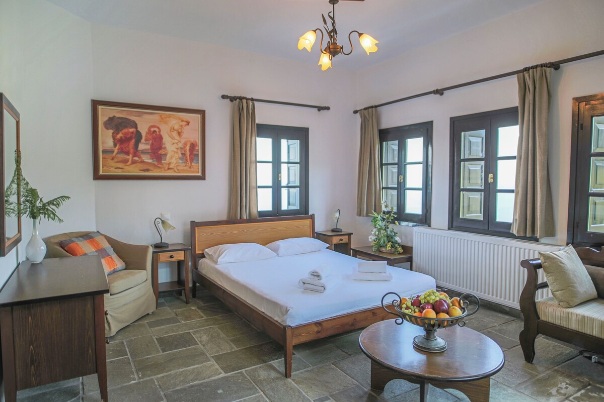 A hotel room with a bed, a bedside table with a lamp, a chair, a sofa, a dining table with fruit, and a painting on the wall.