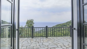 Un balcón abierto con suelo de piedra y barandilla de metal, con vistas a un paisaje montañoso y al mar.