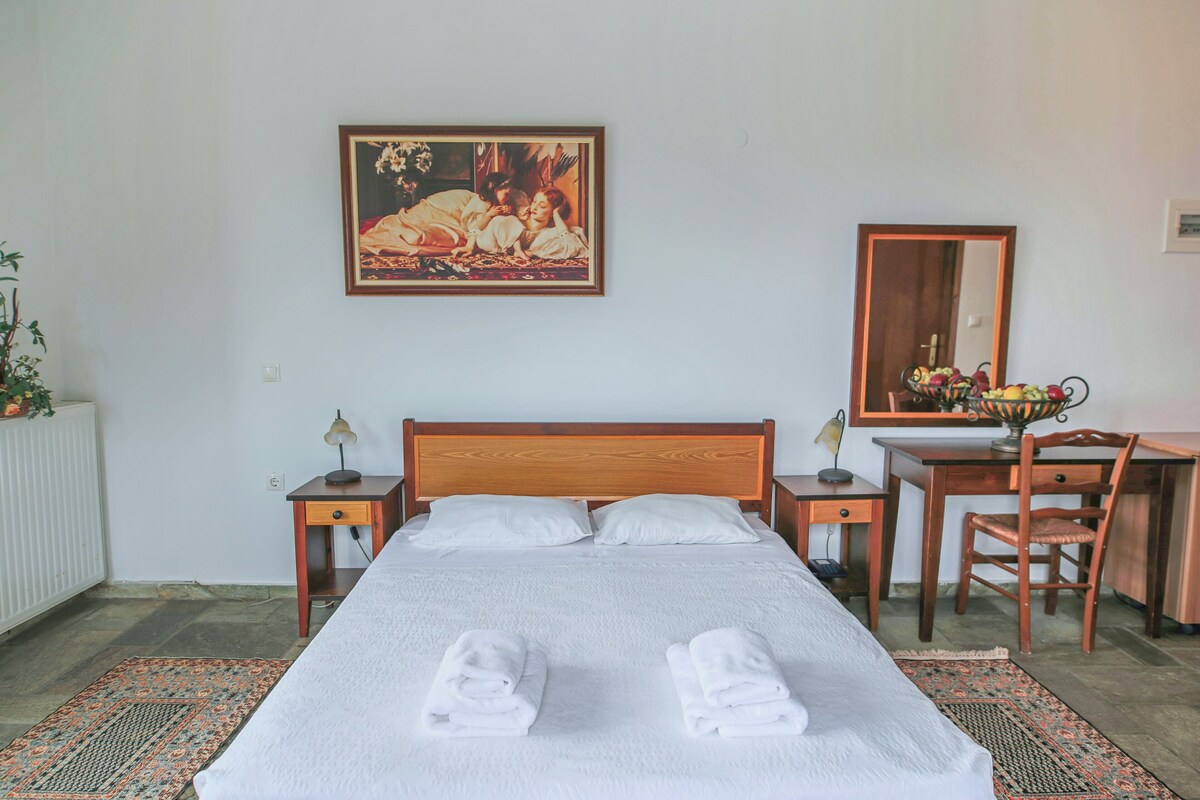 A neatly made bed with two folded towels, a wooden headboard, a painting above the bed, a desk with a chair, and a mirror on the wall.