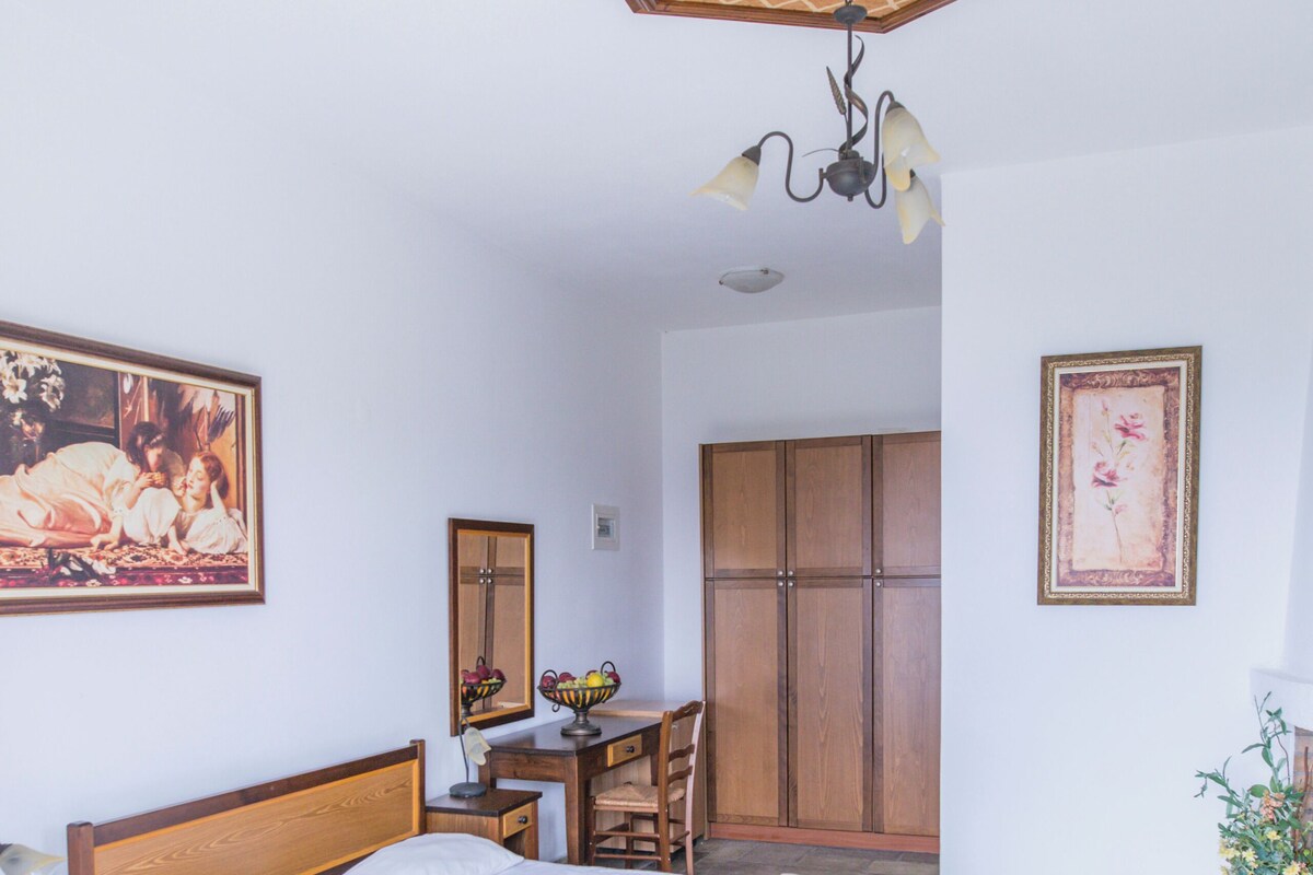 A bedroom with a large bed, wooden wardrobe, a desk with a chair, a mirror, and a ceiling fan.