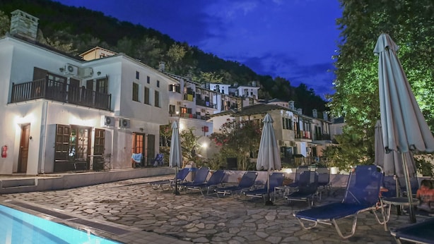 Zona de piscina con sillas de descanso y sombrillas, rodeada de edificios con balcones y suelo empedrado.
