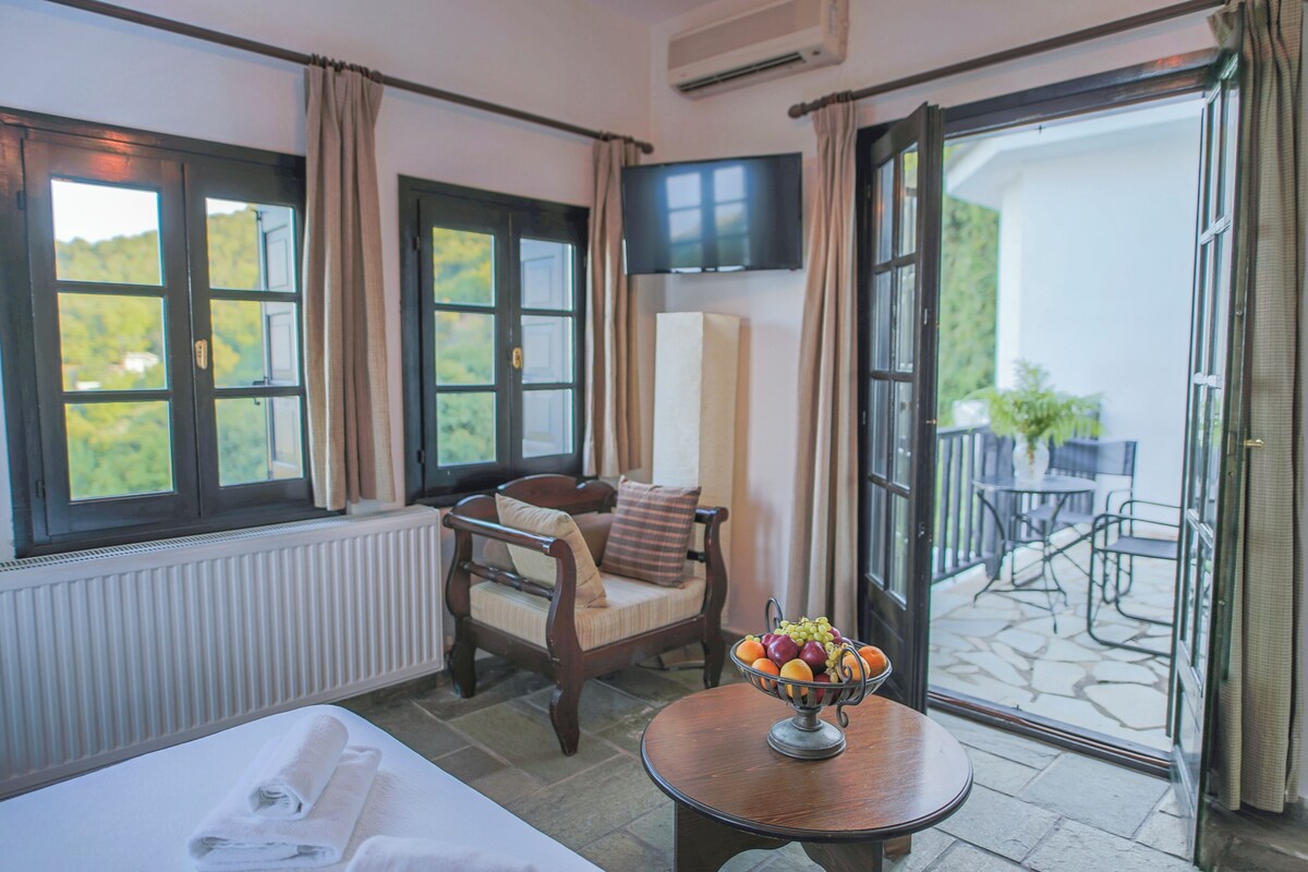 A room with a bed, a chair, a table with fruit, and a view of the outdoors.