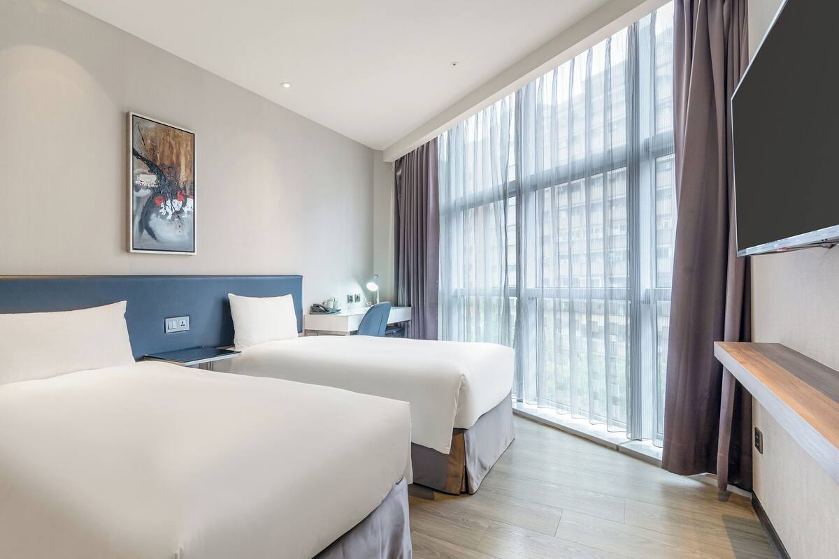 A hotel room with two beds, a desk, a chair, a flat-screen TV, and a large window with curtains.