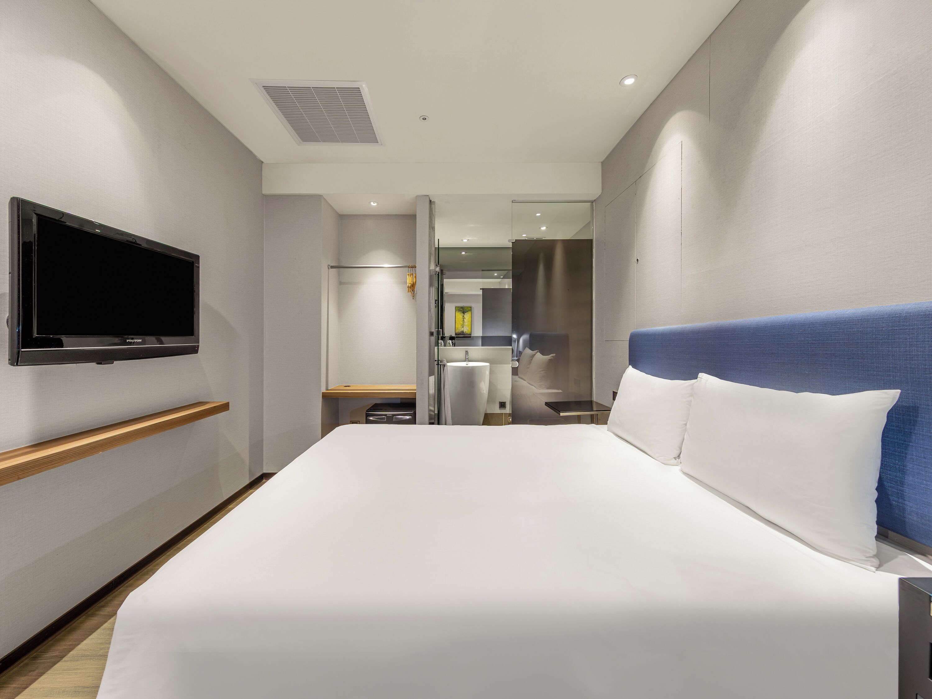 A hotel room with a bed, a television mounted on the wall, a desk, and a bathroom visible through a glass partition.
