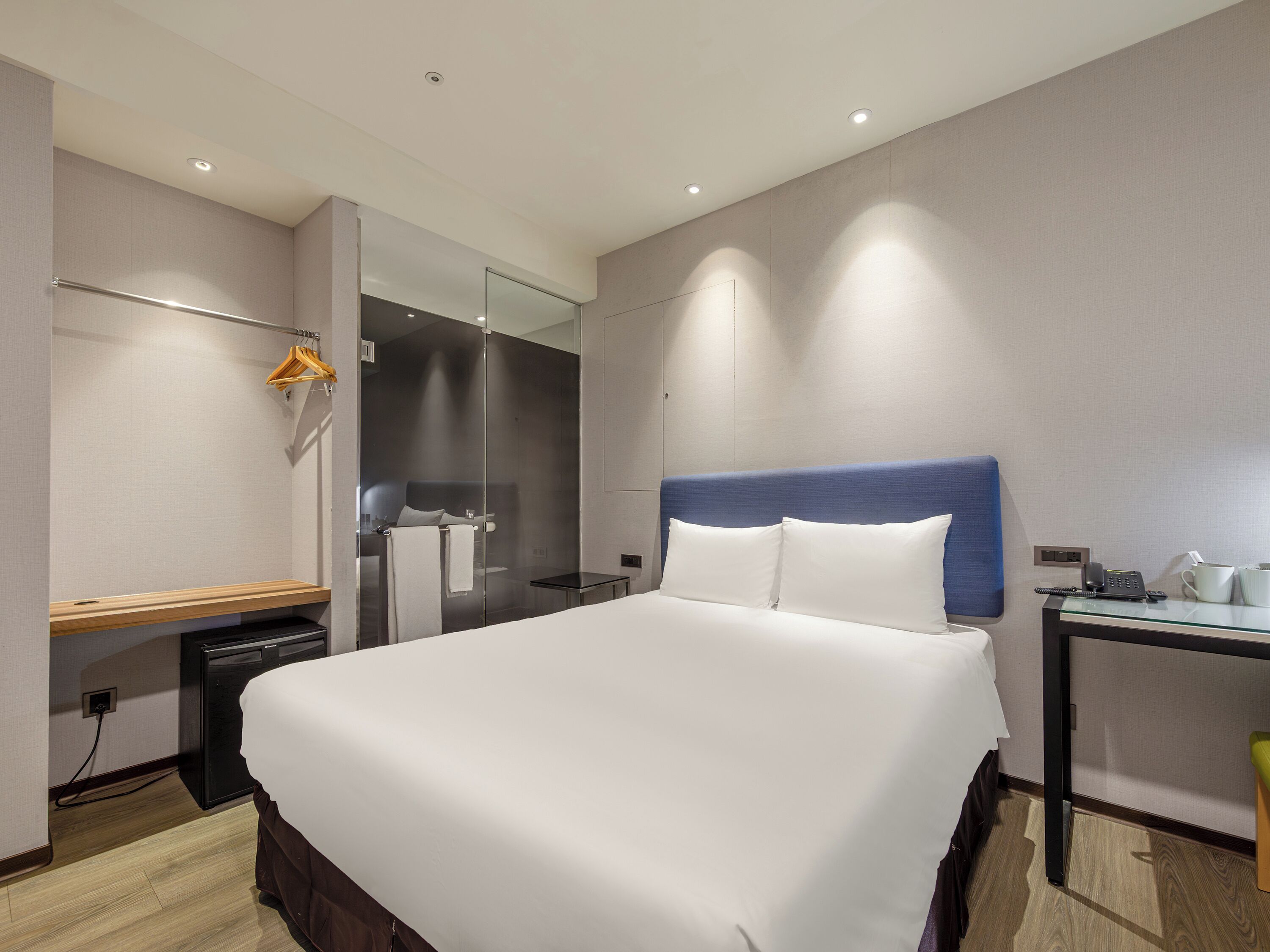 A modern hotel room with a large bed, a bedside table, a mirror, and a small desk with a phone and cup.
