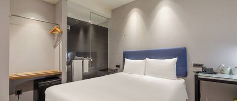 A modern hotel room with a large bed, a bedside table, a mirror, and a small desk with a phone and cup.