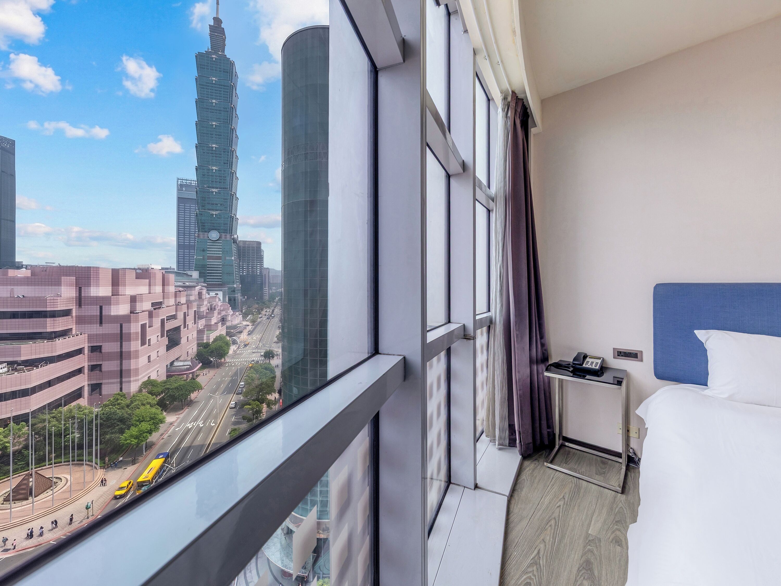 A hotel room with a view of a cityscape, including a tall building and a busy street.
