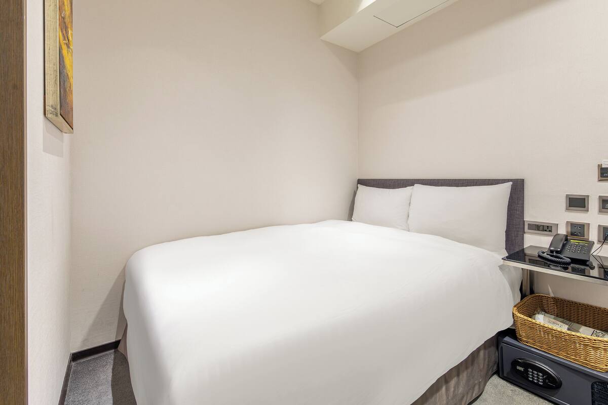 A hotel room with a bed, a bedside table with a phone, a basket, and a wall-mounted control panel.
