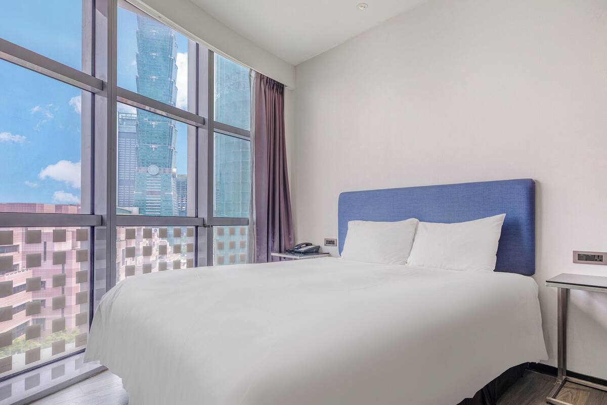 A hotel room with a large window, a bed with white linens, a blue headboard, and a bedside table with a phone.