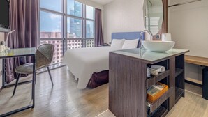 Habitación de hotel con cama, escritorio, silla, ventana grande con vistas a la ciudad y baño con espejo y lavamanos.