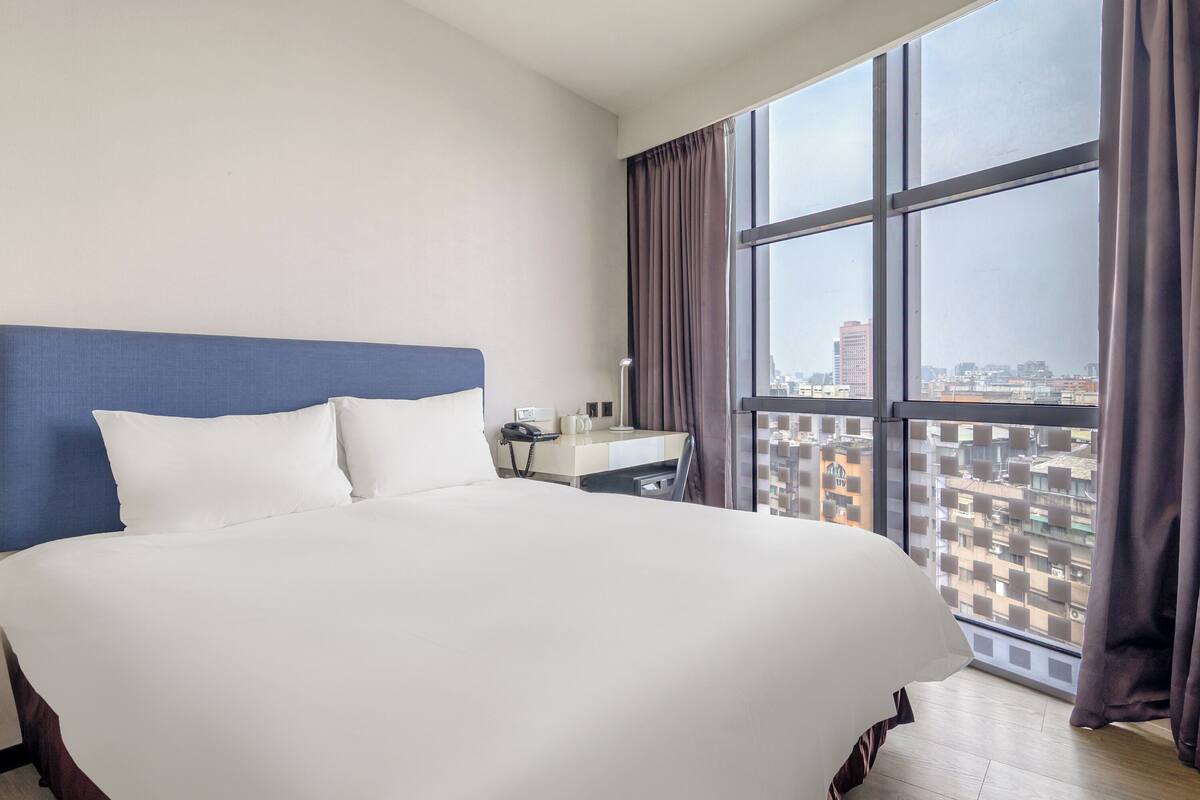 A hotel room with a large bed, a bedside table with a lamp, a phone, and a view of the city through a large window.