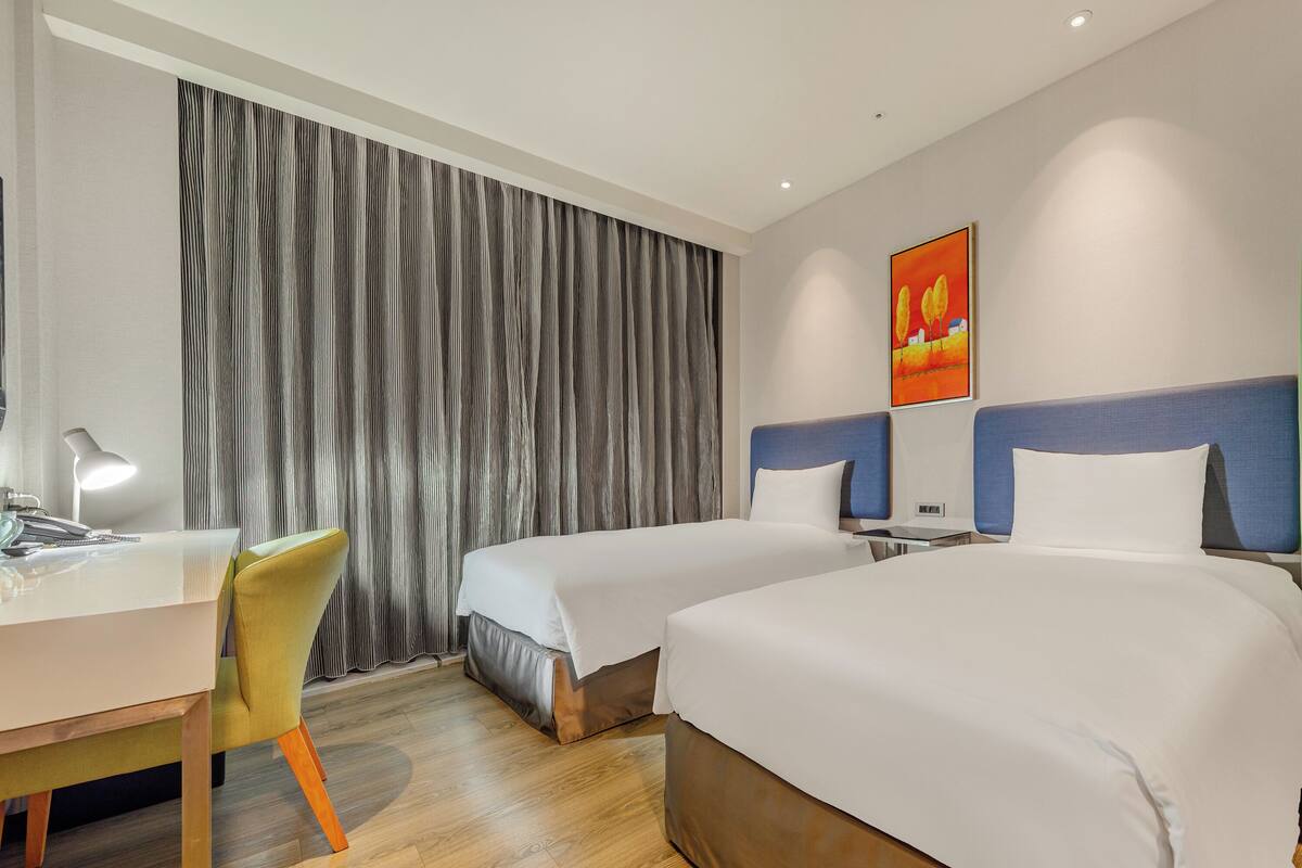 A hotel room with two beds, a desk, a chair, and a large window with curtains.