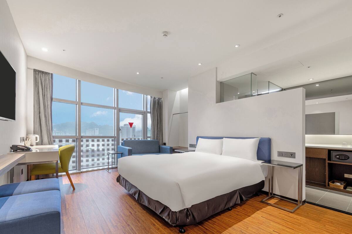 A modern hotel room with a large bed, a desk, a chair, a small table, and a view of the cityscape.